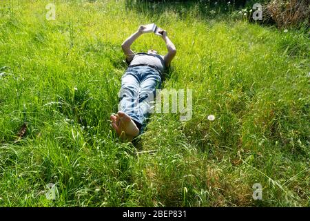 Un uomo legge un libro che giace sull'erba del giardino in primavera durante la distanza sociale per COVID-19 Foto Stock