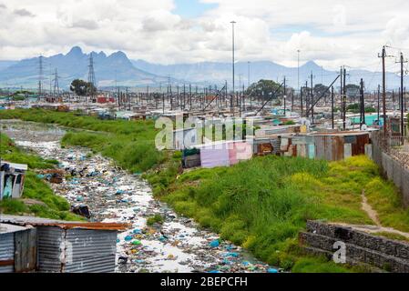 Un fiume è pieno di plastica e altri rifiuti come scorre attraverso una cittadina a Città del Capo. Il fiume inquinato si trova nella baraccopoli di Khayelitsha. L'area contrasta con la lussureggiante campagna sullo sfondo. Non sembra che si sia fatto alcuno sforzo per liberare il fiume. Foto Stock