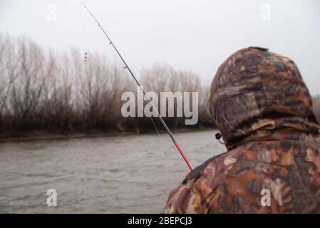 Uomo in camouflage pantaloni e giacca con cappuccio cattura pesce sul fiume in una giornata nuvolosa. Foto Stock