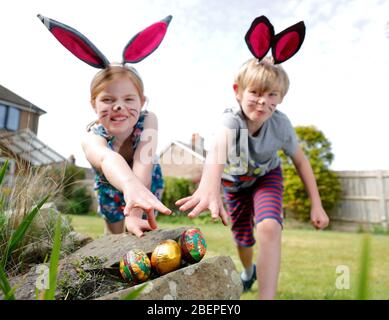 Un fratello e una sorella, indossando la vernice del viso e le orecchie conigliate fatte in casa, prendere parte a una caccia all'uovo di Pasqua nel loro giardino. Foto Stock