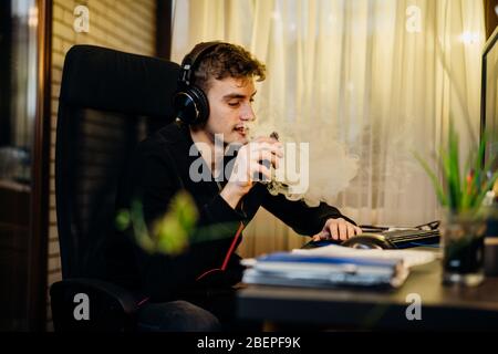 Giovane uomo gamer inalazione di un vapore con sigaretta elettronica.Vape sapore liquido chemicals.Use di e-sigarette in home.Smoking e vaporing negativo Foto Stock