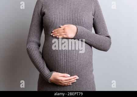 Immagine tagliata di donna incinta in abito grigio che abbraccia il suo grande addome su sfondo grigio. Concetto di maternità. Spazio di copia. Foto Stock