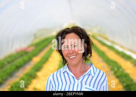 Felice donna ispanica amichevole in piedi in un tunnel agricolo coltivazione fragole sorridente alla macchina fotografica in un ritratto da vicino Foto Stock