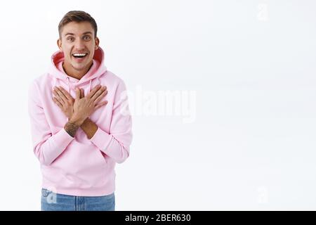 Ritratto di felice biondo ragazzo in felpa con cappuccio rosa, tenere le mani sul petto grato o felice di sentire grande notizia, sorridendo gioire su buona realizzazione Foto Stock