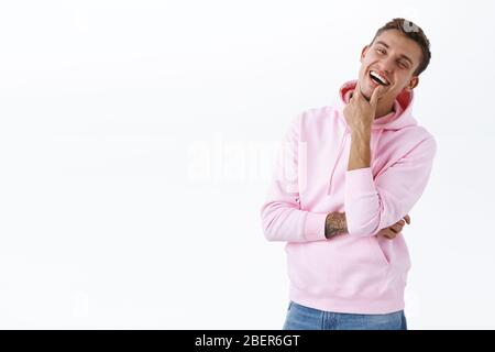 Ritratto di carismatico bel biondo uomo con felpa rosa, ridendo forte, mento touch e fotocamera gazing intrattenuto, guardare film divertente, in piedi Foto Stock