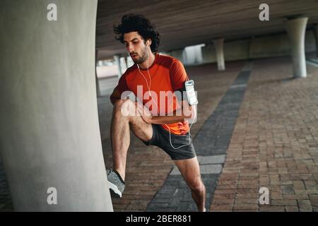 Ritratto di un giovane sano e attraente che ascolta musica sull'auricolare allungando la gamba prima di correre sotto il ponte Foto Stock