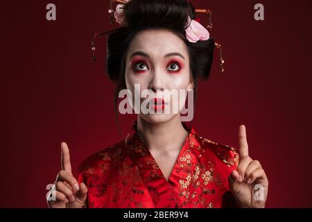 Immagine della giovane donna asiatica geisha in kimono giapponese tradizionale che punta il dito al copyspace isolato su sfondo rosso Foto Stock