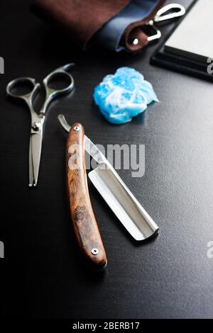Rasoio diritto, schiuma da barba, utensile in pelle e forbici su fondo in legno nero. Attrezzatura per il barbiere Foto Stock