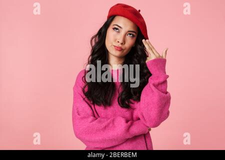 Immagine di seria bella ragazza asiatica che indossa beret gesturing pistola dita al suo tempio isolato su sfondo rosa Foto Stock