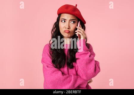 Immagine di dispiaciuti bella ragazza asiatica che indossa beretto accigliato e parlare su smartphone isolato su sfondo rosa Foto Stock