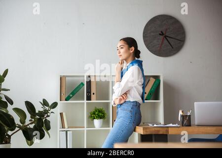 Immagine della giovane segretaria asiatica donna che pensa e sta vicino alla tabella mentre lavora in carica Foto Stock