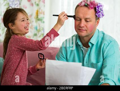 Bambino che gioca e padre disturbare che lavora da casa. Bambina che applica trucco con spazzola. Uomo seduto sul divano con il laptop. Spesa in famiglia Foto Stock