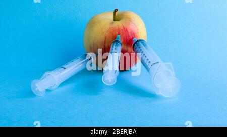Tre siringhe mediche diverse che si attaccano in profondità in una mela su sfondo blu. Il frutto è cosparso di siringhe. Una mela rossa matura traforata da al Foto Stock