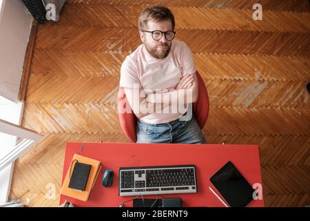 Uomo pensivo su una sedia con le braccia incrociate, lavora sul computer a casa. Concetto di autoisolamento durante l'epidemia di virus. Foto Stock