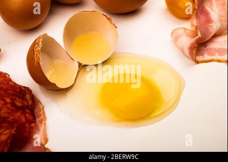 Un uovo di pollo spezzato, fette di salsiccia e pancetta, e uova sparse su sfondo bianco. Primo piano Foto Stock