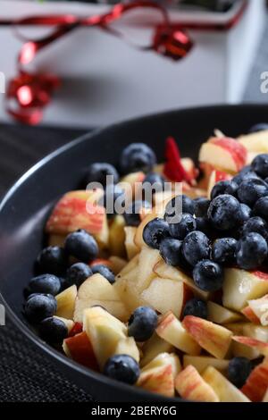 Insalata di frutta su un piatto scuro con bouquet di rose rosse sullo sfondo. Mirtillo, mela, fragola vitamina frutta Foto Stock