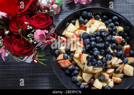 Insalata di frutta su un piatto scuro con bouquet di rose rosse sullo sfondo. Mirtillo, mela, fragola vitamina frutta Foto Stock