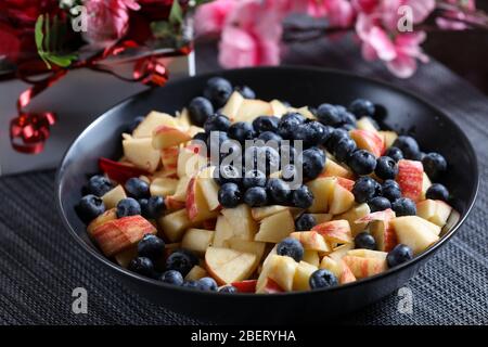 Insalata di frutta su un piatto scuro con bouquet di rose rosse sullo sfondo. Mirtillo, mela, fragola vitamina frutta Foto Stock