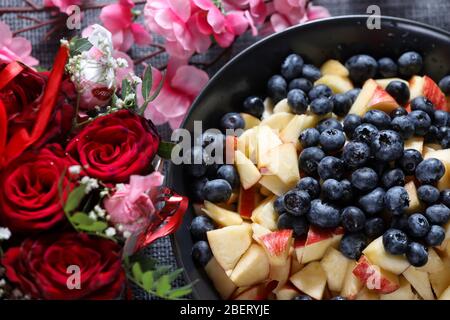 Insalata di frutta su un piatto scuro con bouquet di rose rosse sullo sfondo. Mirtillo, mela, fragola vitamina frutta Foto Stock