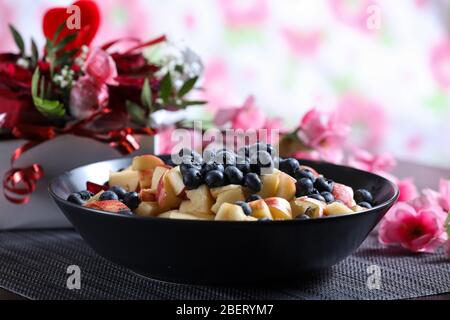Insalata di frutta su un piatto scuro con bouquet di rose rosse sullo sfondo. Mirtillo, mela, fragola vitamina frutta Foto Stock