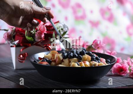 Insalata di frutta su un piatto scuro con bouquet di rose rosse sullo sfondo. Mirtillo, mela, fragola vitamina frutta Foto Stock