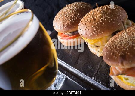 tazza di birra con hamburger e patatine fritte da vicino, bicchieri di birra con birra artigianale scura e leggera con schiuma, su uno sfondo di hamburger, con una varietà Foto Stock
