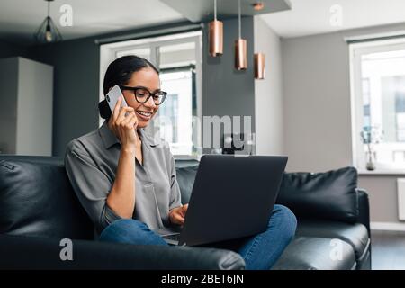 Donna sorridente seduta su un divano con il laptop sulle gambe e facendo una telefonata Foto Stock