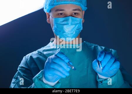 primo piano ritratto di un medico caucasico chirurgo, in un abito sterile, maschera, tenendo un bisturi e porta-ago con un ago, guidandoli all'opera Foto Stock