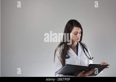 Donna che si occupi di un'assistenza sanitaria che esamina la cartella paziente isolata su sfondo grigio. Stetoscopio intorno al collo, cappotto bianco e bottoni. Foto Stock