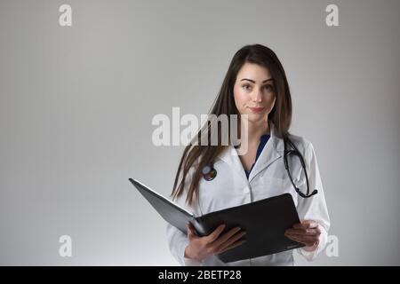 Donna che si occupi di un'assistenza sanitaria che esamina la cartella paziente isolata su sfondo grigio. Stetoscopio intorno al collo, cappotto bianco e bottoni. Foto Stock