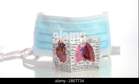 Una maschera chirurgica fotografata insieme ad un diagramma medico cubico dei polmoni e del cuore su sfondo bianco con un riflesso. Foto Stock
