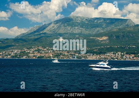 Barche bianche in movimento sull'acqua della baia di Boka Kotor di fronte alla città di Herceg Novi, Monte Orjen della catena montuosa delle Alpi Dinariche, cielo blu con nuvole bianche Foto Stock