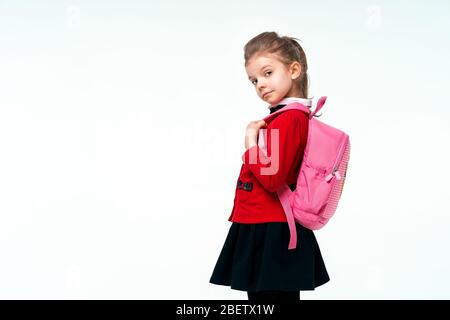 Adorabile bambina in giacca rossa scuola, abito nero, sulle cinghie di uno zaino e sorridente e guardando la macchina fotografica, posa su sfondo bianco studio. Isolare. Foto Stock