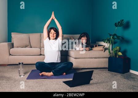 La donna sta facendo yoga online con il laptop durante l'isolamento di sé nel suo soggiorno, senza attrezzi di allenamento, suggerimenti per la meditazione per i principianti. Sua figlia sta leggendo. Famiglia con i bambini, rimanere a casa. Foto Stock