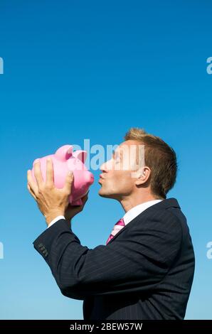 Felice uomo d'affari baciare rosa Piggy banca all'aperto in blu luminoso cielo copia spazio Foto Stock