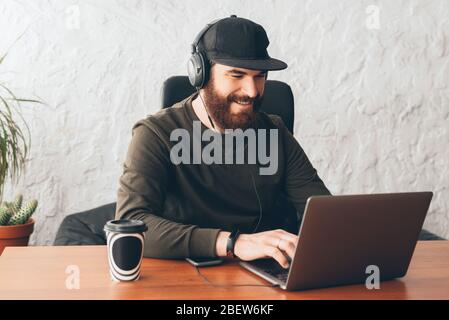 Foto di un giovane con barba sorridente e con le cuffie e lavorando sul laptop in ufficio Foto Stock