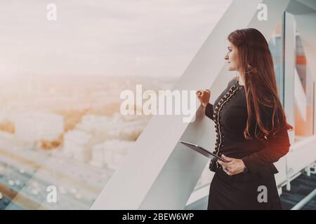Una donna ispanica maturo e sinuosa, imprenditore con un tablet digitale, sta stando in piedi vicino alla finestra di un grattacielo per uffici aziendali e guardando l'autostrada; Foto Stock