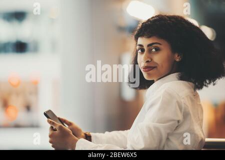 Ritratto indoor di una giovane donna afro-americana scintillante in una camicia bianca con un cellulare e capelli ricci Afro guardando la macchina fotografica; un bira affascinante Foto Stock