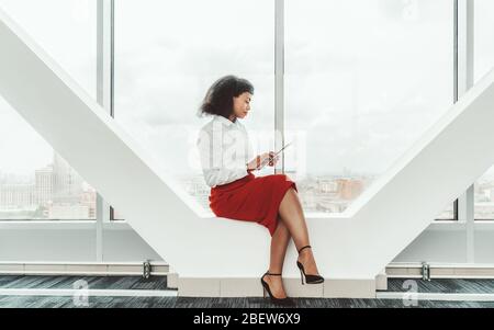 Giovane bella donna d'affari afro-americana con un PC tablet è seduto su un fascio portante di un moderno e luminoso passaggio per ufficio accanto a. Foto Stock