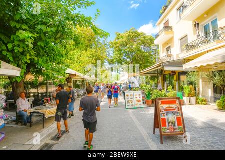 I turisti camminano lungo il percorso affollato passando per caffè, venditori ambulanti e negozi di souvenir nel quartiere Plaka di Atene, Grecia, in una giornata estiva Foto Stock