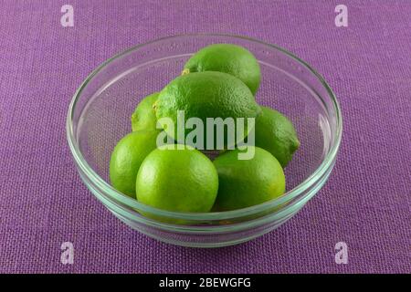 Lime crude fresche in ciotola di frutta di vetro su tovaglia viola Foto Stock