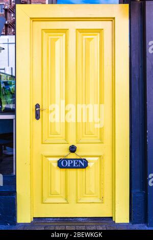 La porta in legno rivestita di giallo brillante con 'APERTO' appeso a una manopola della porta in ottone crea un contrasto accattivante su una strada Launceston in Tasmania. Foto Stock