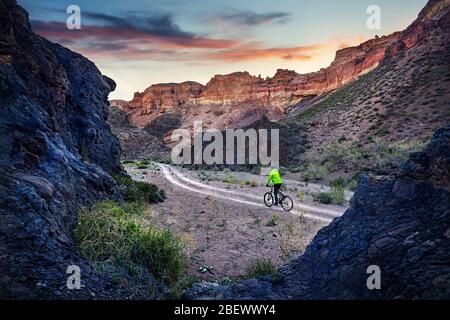 Rider in giacca verde in mountain bike nel deserto Charyn Canyon presso sunrise in Kazakistan. Sport estremo concetto. Foto Stock