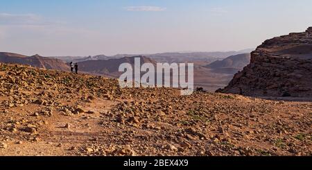 una giovane coppia e un gregge di stambecchi che camminano vicino al tramonto sul bordo del cratere makhtesh ramon in israele con il katum del monte sullo sfondo Foto Stock