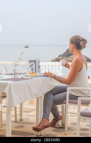 Una donna che beve un caffè con il suo computer portatile seduto in un ristorante vicino alla spiaggia una mattina presto. Foto Stock