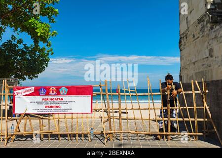 Kuta, Bali Island, Indonesia - 16 aprile 2020: L'uomo di pecalang balinese rimane con il cartello "Rimani a casa" sul cancello d'ingresso alla spiaggia chiuso Foto Stock