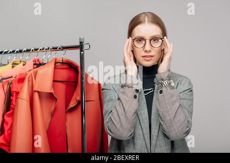 giovane stilista che tocca occhiali vicino abbigliamento alla moda isolato sul grigio Foto Stock