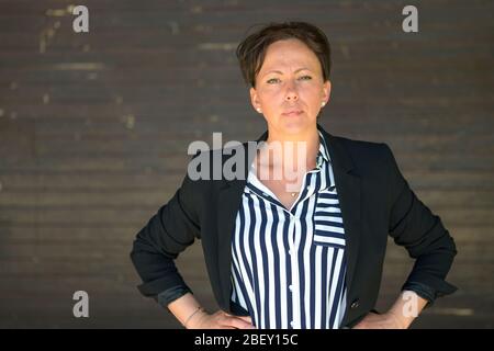 Donna ispanica sicura che si stella con cura alla fotocamera con un'espressione intensa e le mani sui fianchi contro una parete esterna grigia Foto Stock