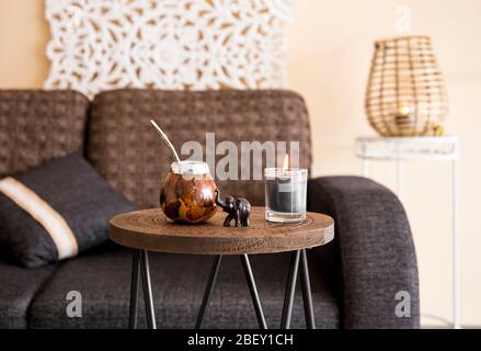 Vista ravvicinata del calabash del tè della yerba mate con paglia di bombilla del metallo e figurina dell'elefante sul piccolo tavolo nel soggiorno moderno, divano sullo sfondo del divano. Foto Stock