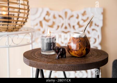 Vista ravvicinata del calabash del tè della yerba mate con paglia di bombilla del metallo e figurina dell'elefante sul piccolo tavolo nel soggiorno moderno, divano sullo sfondo del divano. Foto Stock
